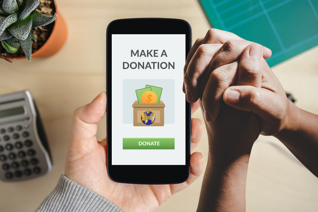 A phone showing a Make a Donation screen, held beside a pair of clasped hands.