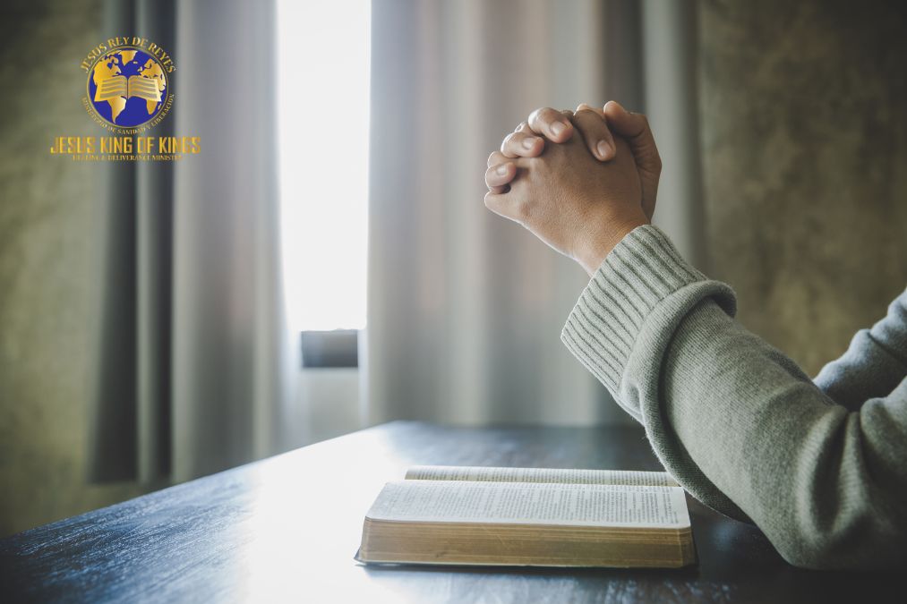 Hands folded in prayer over an open Bible beside a sunlit window.